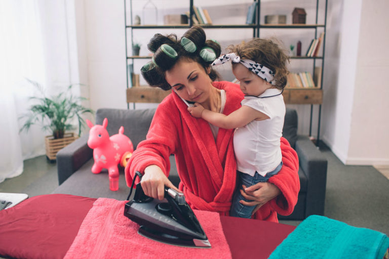 mother and baby together engaged in housework Ironing clothes . Housewife and kid doing homework. Woman with little child in the living room. Homemaker doing many tasks while looks after her infant.